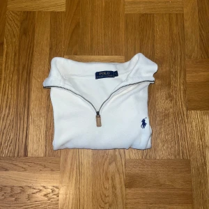 Vit half zip tröja från Polo Ralph Lauren - Vit långärmad tröja från Polo Ralph Lauren med half zip och klassisk krage. Tröjan har blå broderad logga på bröstet och är gjord i mjuk bomull. Perfekt för dig som gillar clean och stilren look.