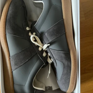 Maison Margiela grå sneakers - Maison Margiela sneakers i grått med beige snörning och gummisula. Ovandelen är en mix av mocka och skinn, med klassisk rund tå och platt sula. Snygg retrostil inspirerad av sportskor från 70-talet. Kommer med originalkartong.