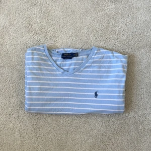 Blåvit randig t-shirt Polo Ralph Lauren - Fräsch blå t-shirt med vita ränder från Polo Ralph Lauren, classic fit. V-ringad hals och korta ärmar, med den ikoniska broderade loggan! Lägg märke till den lilla fläckan på framsidan!