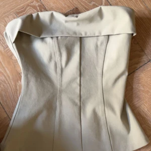 Beige korsett-topp från H&M Divided - Säljer en beige korsett-topp från H&M Divided i storlek XXS. Toppen har en snygg axelbandslös design med bred kant upptill och figurnära passform. Materialet är mjukt och stretchigt, perfekt för att skapa en clean och trendig look.