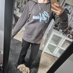Grå hoodie från Hollister med tryck - Säljer en grå hoodie från Hollister med ljusblått 'California'-tryck på bröstet och broderad Hollister-logga vid fickan. Tröjan har huva med snörning, magficka och långa ärmar. Perfekt för chill dagar och sköna outfits.