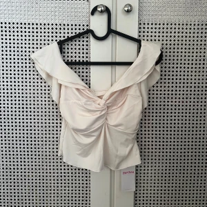 Ljus offshoulder topp från Marthea - Aldrig använd. Supersnygg ljus offshoulder topp från Marthea med volangdetaljer och twistad front. Sitter skönt och har en feminin siluett som ger extra stilpoäng. Perfekt att matcha med jeans eller kjol för en trendig look. Materialet är mjukt och har en lätt glans. 