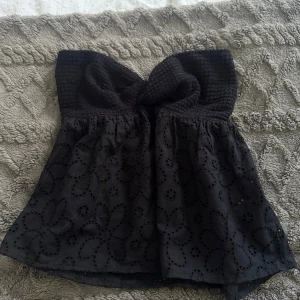 Svart bandeau tubtopp med spetsdetaljer - Säljer en svart bandeau tubtopp med broderade blommor och spetsdetaljer. Toppen har en snygg knut framtill och är axelbandslös, perfekt för dig som vill ha en cool och feminin look. Materialet är mjukt och luftigt med broderad spets.