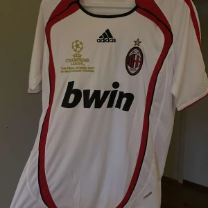 AC Milan Champions League 2007 tröja - AC Milan fotbollströja från Champions League-finalen 2007, officiell Adidas med Bwin sponsortryck. Vit med röda och svarta detaljer, klubbmärke och guldtryck på bröstet. Tillverkad i lätt polyester, kortärmad och sportig passform.