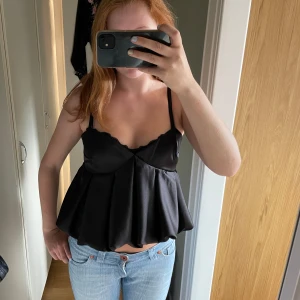 Svart satinlinnetopp med volang - Supersöt svart linnetopp i satin med tunna axelband och volangig, utställd nederdel. Toppen har omlott-detalj framtill och smock i ryggen för extra komfort. Perfekt att styla med jeans eller kjol för en trendig look. Endast använd en gång!