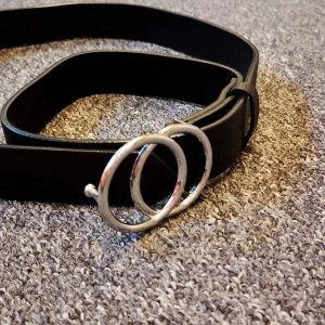 Svart bälte med dubbel ring från H&M - Snyggt svart bälte från H&M Divided med två silverfärgade ringar som spänne. Bältet är stilrent och passar perfekt till jeans eller kjol. Tillverkat i syntetmaterial och har en klassisk, rak form.