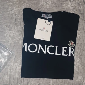 Svart Moncler t-shirt med logga - Svart t-shirt från Moncler i storlek M med stor vit Moncler-logga tryckt på bröstet och en liten broderad patch på vänster sida. Klassisk rund hals och korta ärmar. Materialet är mjuk bomull som känns skönt mot huden. helt ny!