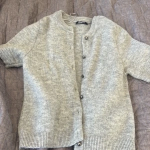 Grå stickad kofta från Gina Tricot - Kortärmad grå stickad kofta från Gina Tricot med rund hals och silvriga knappar framtill. Mjuk och skön känsla med ribbade muddar vid ärmslut och nederkant. Perfekt att slänga över en t-shirt för en chill look.