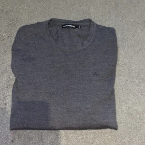 Grå stickad tröja från J.Lindeberg - Snygg grå stickad tröja från J.Lindeberg i storlek M. Tröjan har rund hals och diskret logga på bröstet. Den är långärmad och har ribbade muddar vid ärmslut och nederkant. Perfekt för dig som gillar stilrena och klassiska plagg.