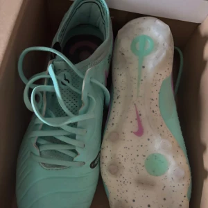Mintgröna Nike fotbollsskor - Säljer ett par mintgröna Nike fotbollsskor med snörning och rosa detaljer. Skorna har en rund tå och platt sula med dobbar för bra grepp på planen. Ovandelen är i syntetmaterial och insidan har ett mönstrat foder. Perfekt för dig som vill sticka ut på fotbollsplanen!