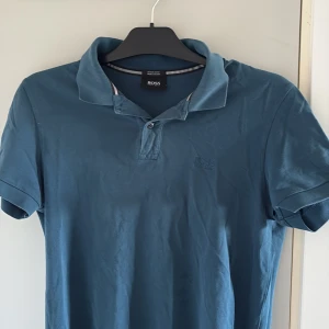 Blå Boss kortärmad pikétröja - STORLEK S-XS (170-178)Snygg blå pikétröja från Hugo Boss i regular fit, tillverkad av mjuk pima bomull. Klassisk krage med två knappar och diskret Boss-logga broderad på bröstet. Perfekt för dig som gillar stilrena och bekväma plagg.