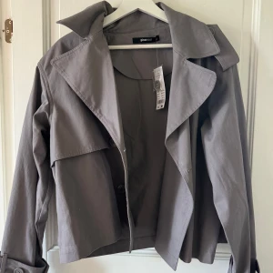 Grå croppad trenchcoat från Gina Tricot - Snygg croppad trenchcoat i grått från Gina Tricot med bred krage och knappar framtill. Jackan har en cool, boxig passform och långa ärmar. Perfekt för dig som gillar stilrena och trendiga plagg med lite edge.