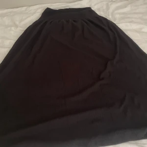 Svart långkjol från Gina Tricot - Svart långkjol från Gina Tricot med smockad midja som ger en skön och flexibel passform. Kjolen är enkel och stilren utan mönster, tillverkad i ett mjukt syntetmaterial. Perfekt för dig som gillar minimalistisk och bekväm stil.