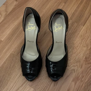 Svarta klackar från Christian Louboutin - Säljer ett par svarta klackar från Christian Louboutin med ikonisk röd sula. Skorna har öppen tå, rundad form och snygga veckade detaljer framtill. Tillverkade i blankt skinn som ger en lyxig vibe. Perfekt för dig som vill sticka ut med exklusiva Paris-vibes.