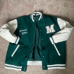 Grön och vit collegejacka Divided L - Collegejacka från Divided i storlek L, mörkgrön med vita ärmar och detaljer. Broderad text 'Minnesota College' och stor M på bröstet. Jackan har ribbade muddar, knappar framtill och två fickor. Materialet är polyester och jackan har en klassisk varsitystil.