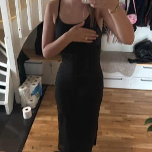 Svart långklänning med smala axelband - Snygg svart långklänning med smala axelband och figurnära passform. Klänningen är ärmlös och har en enkel, stilren design som ger en cool och clean look. Perfekt för dig som gillar minimalistiskt och tidlöst mode.