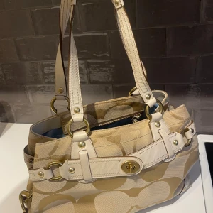Beige handväska från Coach - Snygg beige handväska från Coach. Väskan har guldfärgade metalldelar och dubbla handtag. Insidan är fodrad med ljusblått tyg och har en innerficka med dragkedja. Väskan är äkta, köpt i Japan för 6 månader sedan. Har några fläckar på insidan men i  fint skick annars! 