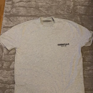 Grå Essentials Fear of God t-shirt - Snygg grå t-shirt från Essentials Fear of God ( OverSized M )  med svart logotryck på bröstet och stort tryck på ryggen. Klassisk rund hals och korta ärmar. Perfekt för dig som gillar streetwear och enkel stil. Materialet känns mjukt och skönt mot huden. 