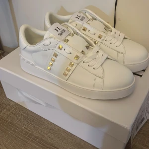 Valentino Garavani Rockstud sneakers - Snygga vita Valentino Garavani Rockstud sneakers med guldiga nitar och klassisk snörning. Skorna har en rund tå och platt sula med mönstrad undersida. Tillverkade i lyxigt skinn och har coola detaljer som ger en exklusiv vibe. Perfekta för dig som gillar statement pieces.Allt originalet finns kvar!
