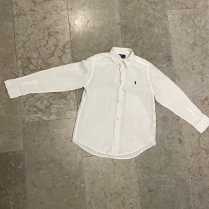 Vit linneskjorta från Polo Ralph Lauren - Vit skjorta från Polo Ralph Lauren i luftig linne, klassisk krage och långärmad modell. Diskret blå logga broderad på bröstet och snygga pärlemorknappar framtill. Perfekt för en clean och fräsch look. Det finns dock en liten missfärgning på ena ärmen men är annars i väldigt fint skick.