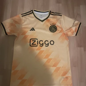 Ajax gul fotbollströja Adidas M - Ajax fotbollströja i gul och orange toner från Adidas, storlek M. Tröjan har korta ärmar, svart-vita detaljer och Ajax-logga med tre stjärnor på bröstet. Ziggo och GigaNet som sponsorer, tillverkad i lätt polyester med geometriskt mönster.