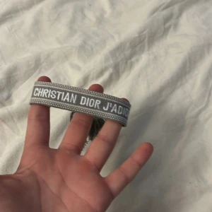 Grått Dior armband - Snyggt grått armband från Christian Dior med texten 'J'Adior' broderad i vitt. Armbandet har flätade band med tofsar i ändarna och är gjort i ett mjukt tygmaterial. Perfekt accessoar för att lyfta din outfit med lite lyxig vibe.