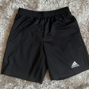 Svarta träningsshorts från Adidas - Säljer ett par svarta träningsshorts från Adidas med elastisk midja och Climacool-teknologi för optimal ventilation. Perfekta för sport och träning. Adidas-logga i vitt på benet.