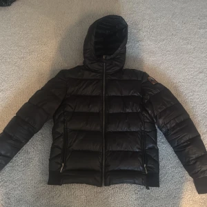 Svart dunjacka från Parajumpers - Säljer en snygg svart dunjacka från Parajumpers med huva och dragkedja. Jackan har en quiltad design och en logotyp på ärmen. Perfekt för kyliga dagar!