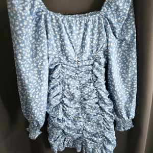 Blå blommig klänning  - Charmig blå klänning med vita blommor och rynkdetaljer framtill. Den har långa ärmar med elastiska muddar och en smickrande passform. Perfekt för en somrig look! Köpt för 250 på shein(använd vid ett tillfälle)