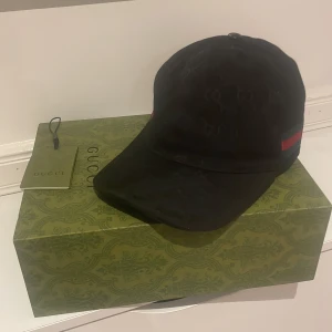 Gucci keps - Snygg svart keps från Gucci med diskret mönster och klassiska röd-gröna detaljer på sidan. Kepsen har en justerbar rem baktill för perfekt passform. Perfekt för att ge en lyxig touch till din outfit. Kommer med påse,box och tagen. Släpper kepsen för 700 utan något med bara kepsen.