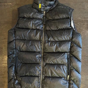Svart dunväst från Parajumpers - Säljer en svart dunväst från Parajumpers med dragkedja och hög krage. Västen har en quiltad design och två fickor med dragkedja framtill. Perfekt för kyligare dagar.