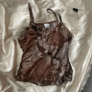 Brun mönstrad linnetopp  - Snygg brun linnetopp med ett unikt mönster som kombinerar leopard och rutigt. Toppen har en v-ringad design och smala axelband, samt paljetter som detaljer som ger en extra touch. Perfekt för att sticka ut! (obs att paljetterna kan kännas mot huden ifall man tycker sånt är jobbigt) 