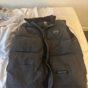 Grå dunväst från Canada Goose - Säljer en stilren grå dunväst från Canada Goose. Västen har en dragkedja och knappar framtill samt två praktiska fickor. Perfekt för kyligare dagar. Tar emot byten också. Såklart äkta