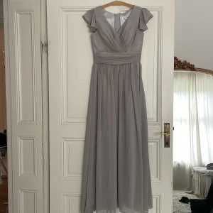 Grå långklänning med volangärmar - Elegant grå långklänning med v-ringad omlott-design och volangärmar. Klänningen har en dragkedja i ryggen och ett brett band i midjan som ger en smickrande passform. Perfekt för speciella tillfällen. Perfekt för bal eller brudtärna 