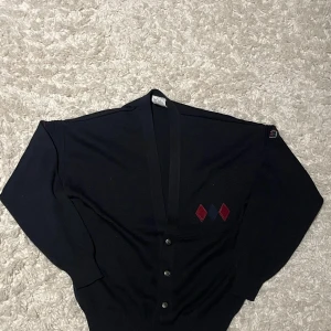 Svart kofta från Herr Collection - Snygg svart kofta från Herr Collection med knappar framtill och dekorativa röda och blå detaljer på bröstet. Perfekt för en stilren look. Koftan har långa ärmar och en klassisk design.