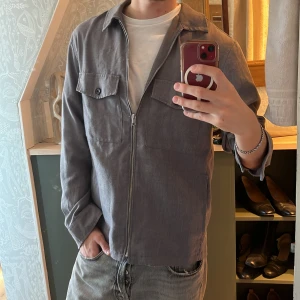 Grå overshirt från H&M - Säljer en stilren grå overshirt från H&M i storlek M. Jackan har en dragkedja framtill och två bröstfickor med knappar. Perfekt för lager-på-lager-stil och passar bra till både jeans och chinos.