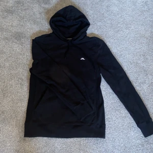 Svart hoodie från J.Lindeberg - Säljer en stilren svart hoodie från J.Lindeberg. Den har en klassisk design med en liten logotyp på bröstet och justerbar huva med dragsko. Perfekt för en avslappnad look.