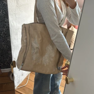 Beige axelväska med strassdetaljer - Säljer en snygg beige axelväska med glittriga strassdetaljer i form av vingar på framsidan. Väskan har dragkedjor på sidorna och är tillverkad i ett mjukt material som liknar skinn. Perfekt för att ge din outfit en extra touch av stil.