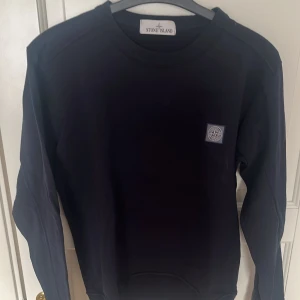 Marinblå tröja från Stone Island - Säljer en stilren svart tröja från Stone Island med deras ikoniska logga på bröstet. Tröjan har långa ärmar och en klassisk rund halsringning. Perfekt för en avslappnad och trendig look.