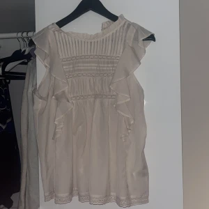 Beige blus med volangdetaljer - Säljer en söt beige blus med volangdetaljer och spets. Blusen är ärmlös och har en romantisk stil med fina detaljer framtill. Perfekt för en somrig look!