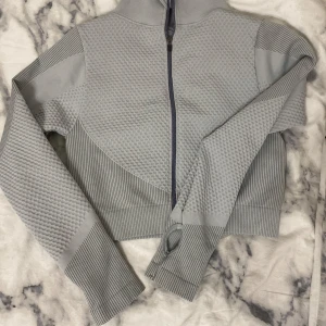 Grå ribbad hoodie med dragkedja - Säljer en grå ribbad hoodie med dragkedja och hög krage. Tröjan är croppad och har långa ärmar med tumhål för extra komfort. Perfekt för en avslappnad stil.