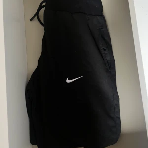 Svarta träningsbyxor från Nike - Snygga svarta träningsbyxor från Nike med en vit logga på benet. Byxorna har en elastisk midja med dragsko för justerbar passform och praktiska sidofickor. Perfekta för sport och träning.