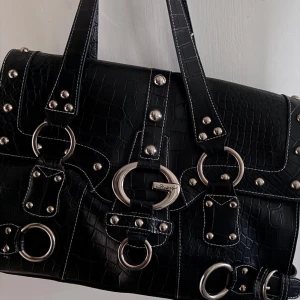 Svart axelväska med nitar - Snygg svart axelväska i skinnimitation med krokodilpräglat mönster. Väskan har flera dekorativa metallringar och nitar