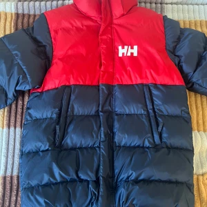Röd och marinblå dunjacka från Helly Hansen - Säljer en snygg och varm dunjacka från Helly Hansen i rött och marinblått. Storlek 164/14 years eller storlek S. Jackan har en hög krage och dragkedja framtill. Perfekt för kalla dagar med sin pufferdesign som håller dig varm. Helly Hansen-loggan syns både fram och bak.