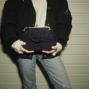 Säljer en snygg mörkblå stickad handväska med guldfärgade detaljer. Väskan har en klassisk ramöppning och en kort handrem. Perfekt för att ge en stilfull touch till din outfit.