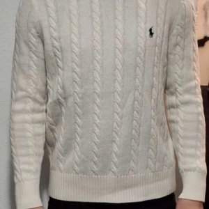 Vit stickad tröja från Ralph Lauren - Säljer en stilren vit stickad tröja från Ralph Lauren med kabelstickat mönster. Tröjan har långa ärmar och en liten broderad logga på bröstet. Perfekt för kyliga dagar!