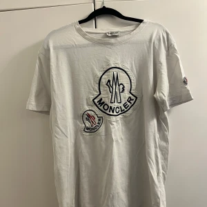 Vit t-shirt från Moncler - Säljer en vit t-shirt från Moncler med stora broderade logotyper på framsidan och en mindre på ärmen. T-shirten har en klassisk rund hals och korta ärmar. Perfekt för en stilren och avslappnad look.