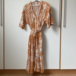 Orange mönstrad klänning från Desmond & Dempsey x H&M - Härlig orange sommarklänning med vitt tropiskt mönster från Desmond & Dempsey x H&M. Klänningen är v-ringad, har armbågslånga vida ärmar och ett knytband i midjan med tofsar. En av tofsarna saknas. Fina klädda knappar framtill. Insydda vita dekorband. Längd 120 cm. Storlek S men passar mig som är ca 40/42. 