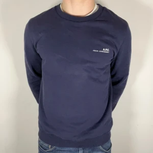 Mörkblå tröja från A.P.C. - Säljer en stilren mörkblå tröja från A.P.C. med tryck på bröstet. Tröjan har en klassisk rund hals och långa ärmar, perfekt för en avslappnad look. Tillverkad i Portugal. Skickat är som nytt, och jag är 178 74 cm 