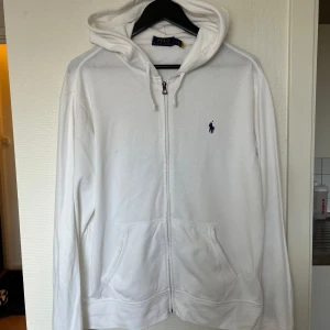 Vit hoodie från Ralph Lauren - Säljer en stilren, tunn vit hoodie från Ralph Lauren med dragkedja och klassisk logga på bröstet. Perfekt för en avslappnad look. Den har långa ärmar och en bekväm huva. Passar bra till både jeans och joggers.